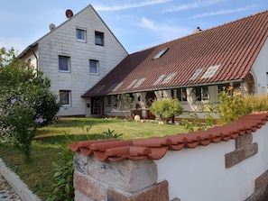 A two-story house with a red-tiled roof, a small porch, and a garden with flowers and shrubs.