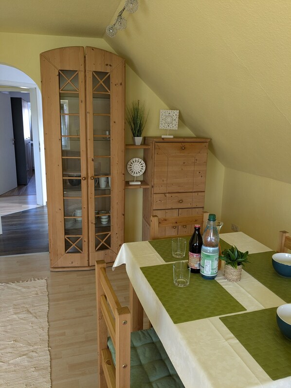 A dining area with a table set for two, a wooden cabinet with glass doors, and a plant on the wall.
