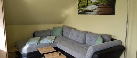 Una sala de estar moderna con un sofá gris, dos mesitas de noche de madera, una lámpara de pie y un cuadro con un paisaje boscoso.