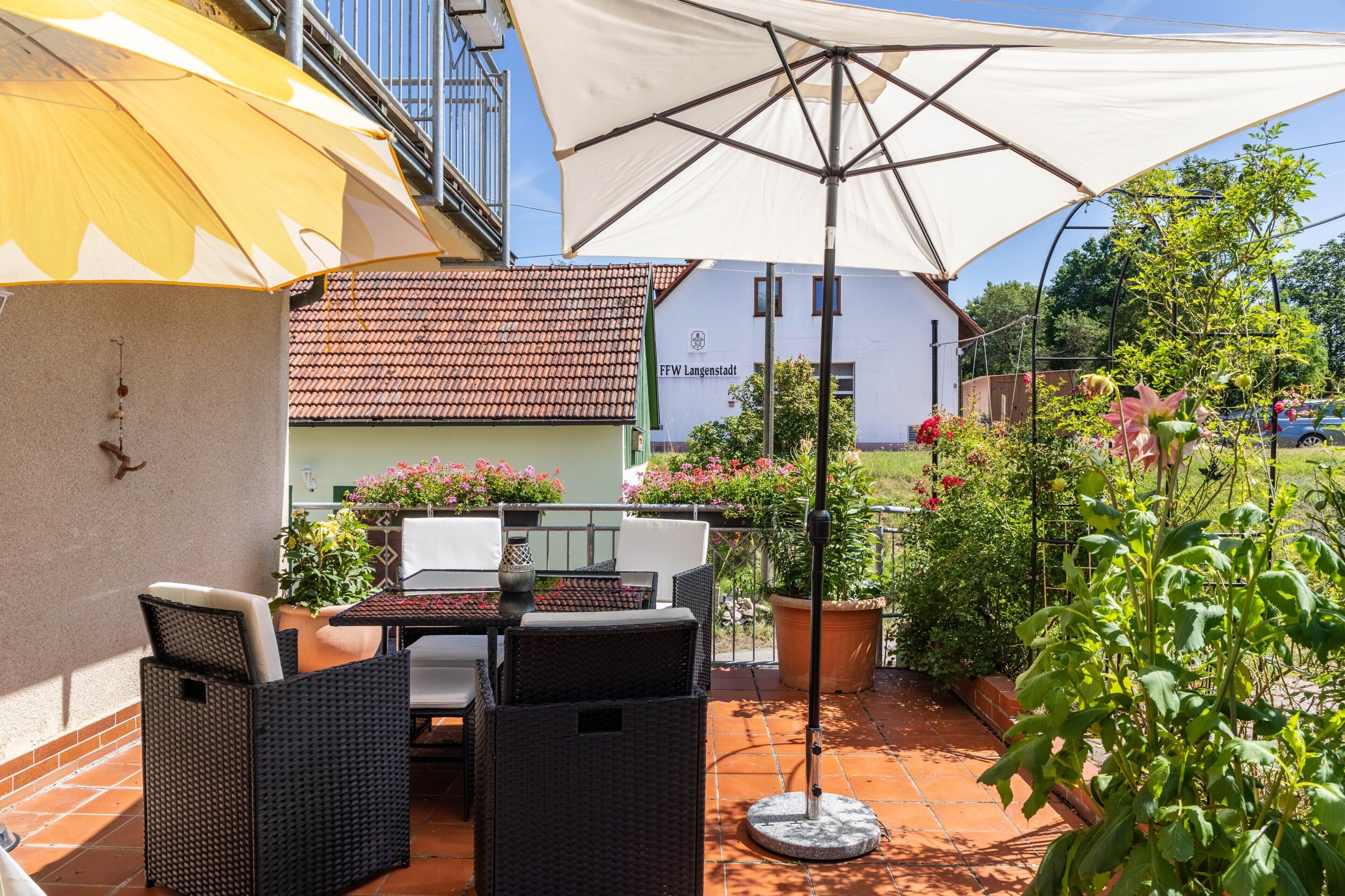 Een terras buiten met rieten meubilair, een grote parasol en potplanten.