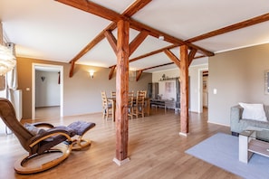 Ampla sala de estar com vigas de madeira, mesa de jantar com cadeiras, sofá e uma poltrona de balanço.