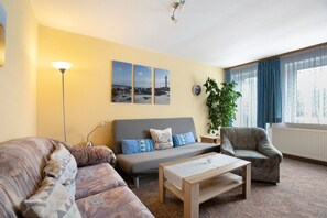 Ein Wohnzimmer mit Sofa, Sessel, Couchtisch und einer Lampe. An der Wand hängen drei gerahmte Bilder, ein Topf mit einer Pflanze und ein Fenster mit Vorhängen.