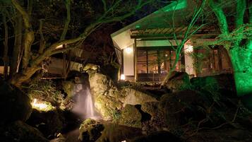 A serene nighttime garden with a waterfall, illuminated by soft lighting, and a modern building with large windows in the background.