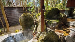 Un área de aguas termales con una terraza de madera, rocas grandes cubiertas de musgo y un banco de madera.