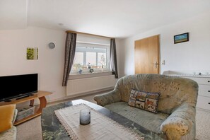 A living room with a sofa, a television, a window with curtains, a clock, and a small picture on the wall. - Apartment 'Plüschmors' with Shared Garden, Balcony and Wi-Fi (Hedwigenkoog)