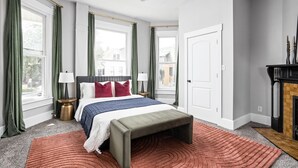 Une chambre à coucher comprenant un lit, un banc, deux tables de chevet, une lampe, une cheminée et de grandes fenêtres avec des rideaux.