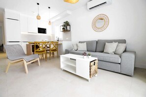 A modern living room with a grey sofa, a white coffee table, and a grey armchair. There is a kitchen area with a dining table and chairs, and a small plant on a shelf.
