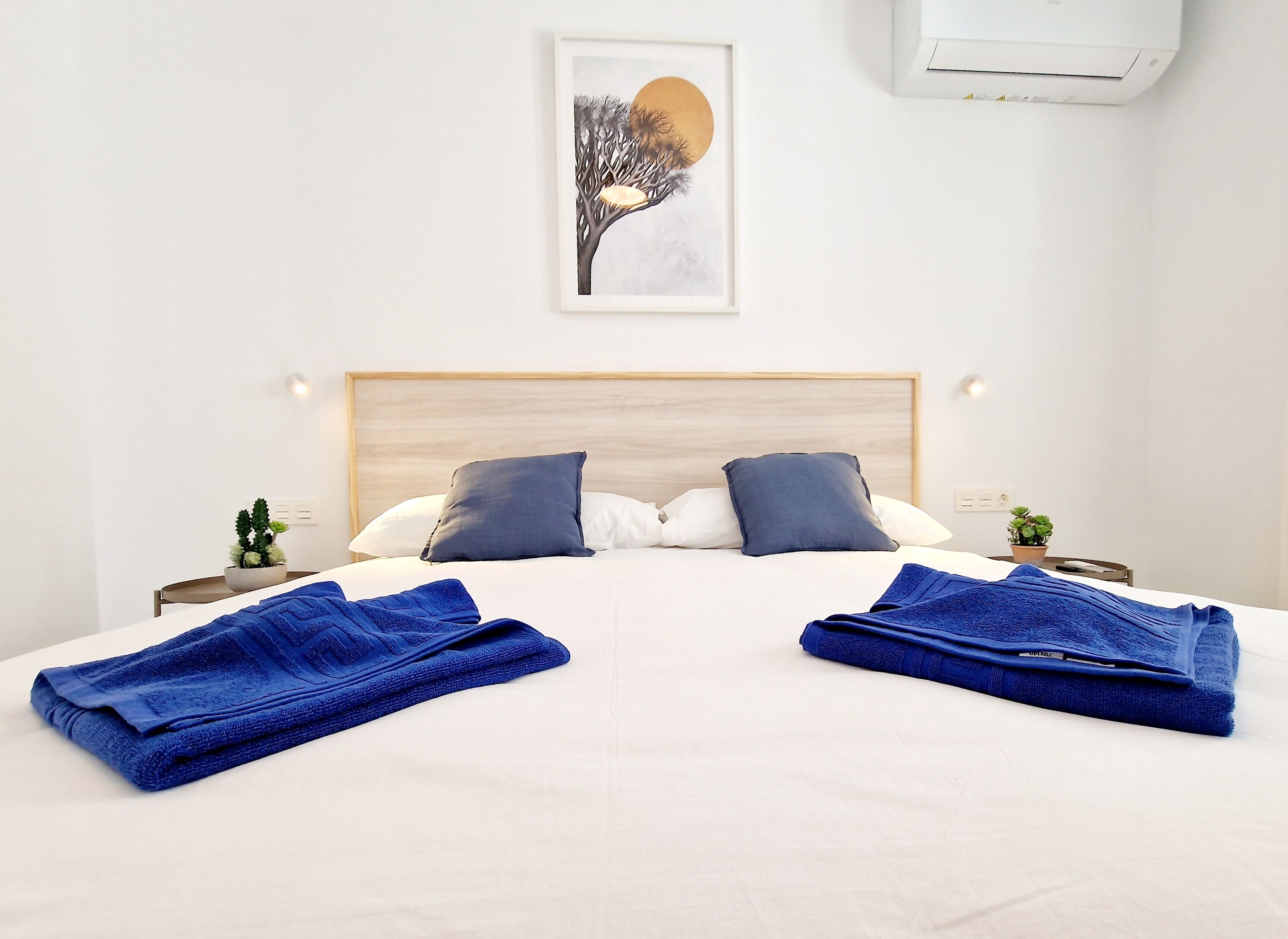 Una cama bien hecha con dos almohadas, una obra enmarcada encima y una planta pequeña en la mesita de noche.