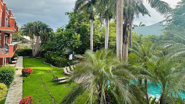 A lush garden with palm trees, a swimming pool, and a red building with balconies.