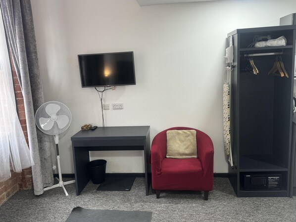 A compact room with a desk, a chair, a TV, a fan, a small trash bin, and a wardrobe.