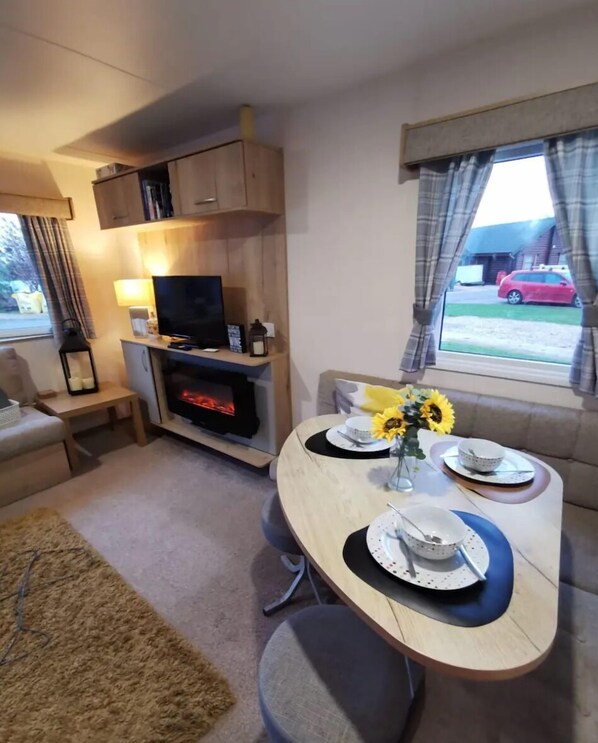 A cozy living area with a TV, fireplace, and dining table set with plates and a vase of sunflowers. - Cosy hottub caravan Retreat in Lincolnshire (Lincoln)
