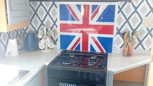 A kitchen with a gas stove, oven, and a Union Jack backsplash. There are cabinets, a countertop, and various kitchen utensils.