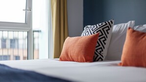Uma cama com almofadas padronadas em branco e preto, um cobertor azul escuro e vista de um prédio através de uma janela com cortinas amarelas.