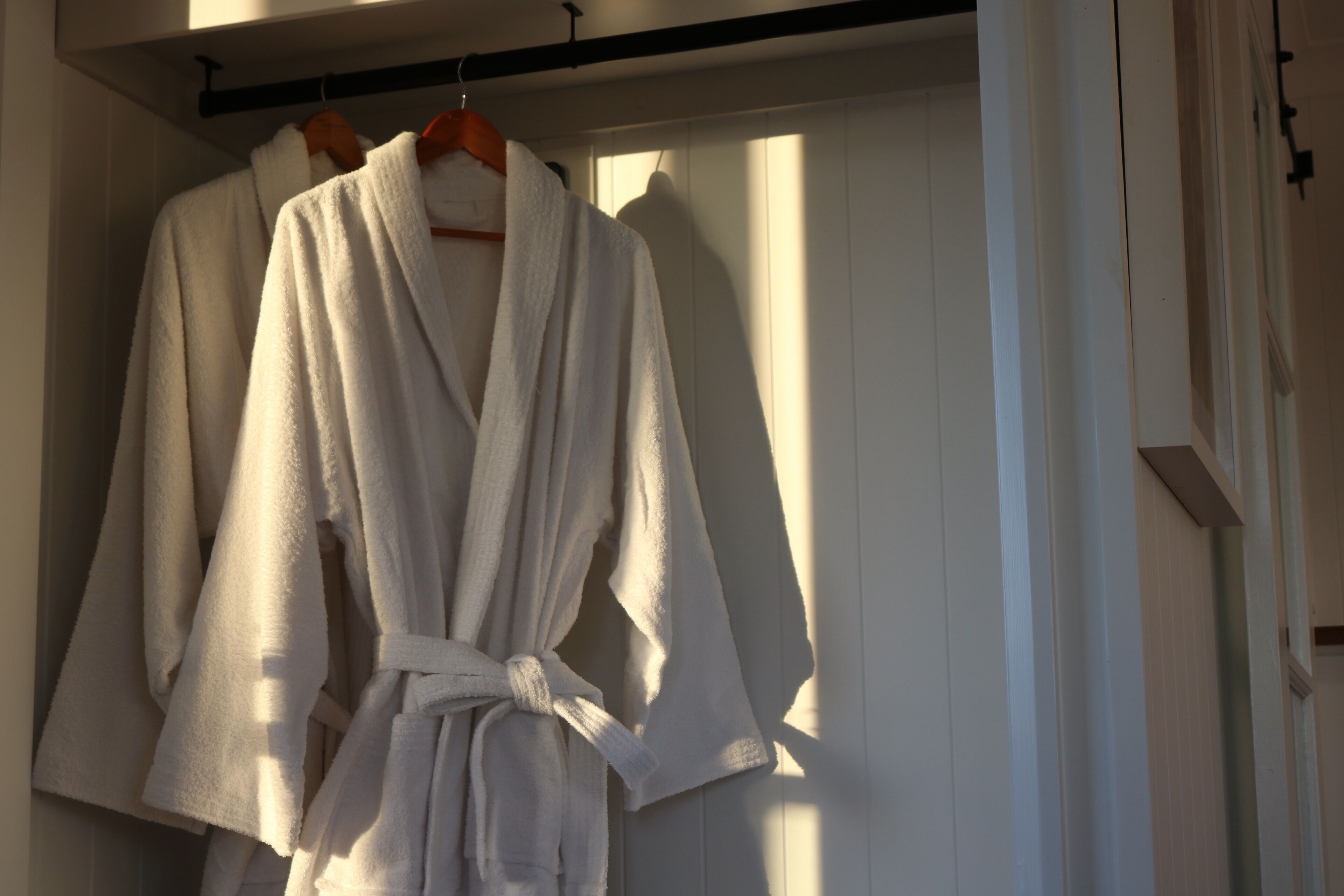 A white robe hanging on a wooden hanger in a room with white walls and a window.