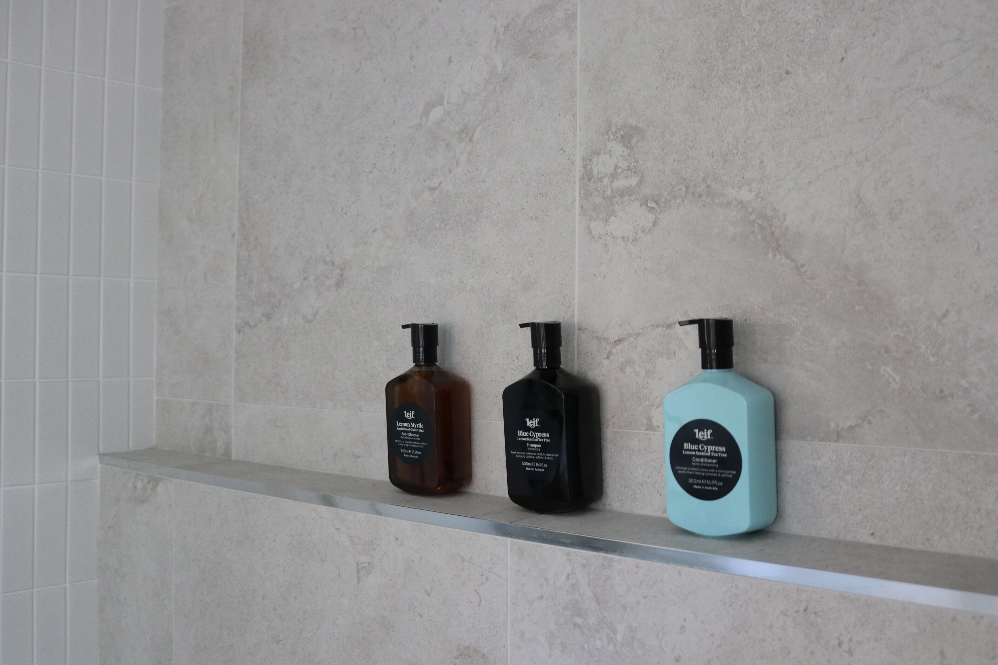 Tres botellas de productos de cuidado personal sobre una estantería frente a una pared de azulejos.