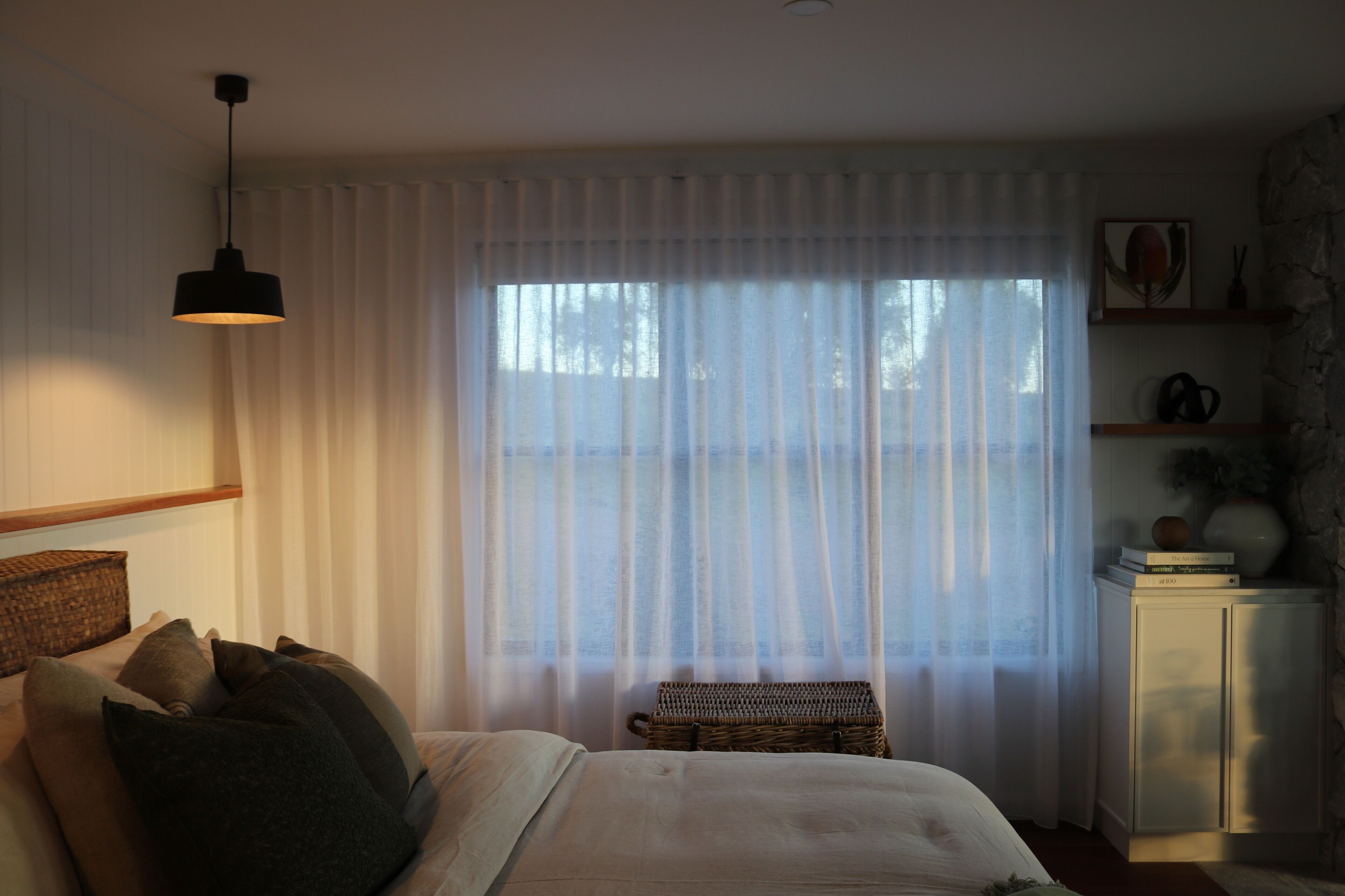 A bedroom with a bed, a hanging light, a window with sheer curtains, a wicker bench, and a cabinet with decorative items.