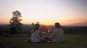 Twee personen zitten op een deken buiten tijdens de zonsondergang, met een boom en een houten constructie op de achtergrond.