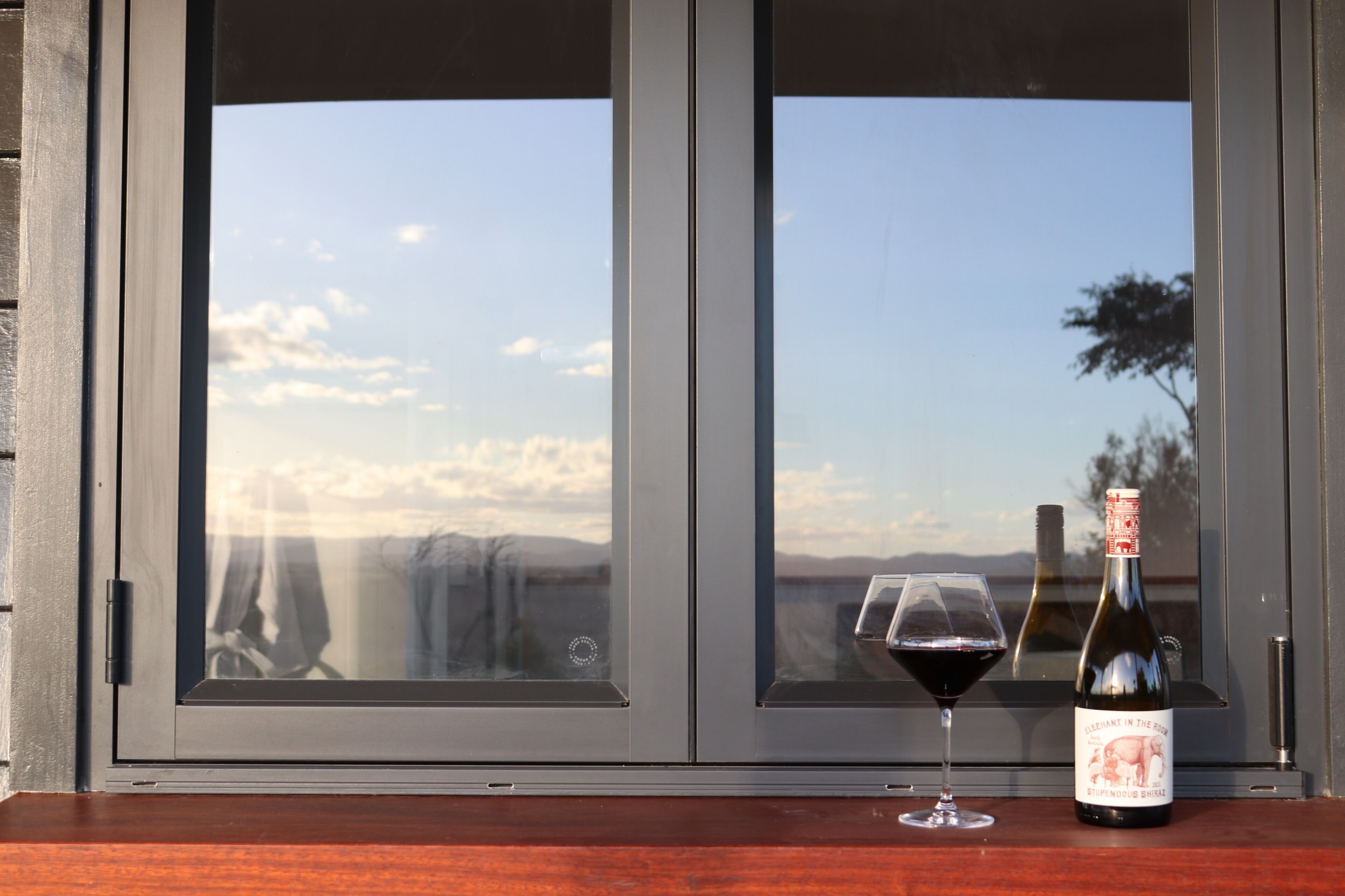 Une bouteille de vin et un verre posés sur une surface en bois, devant une fenêtre donnant sur le ciel et un paysage lointain.