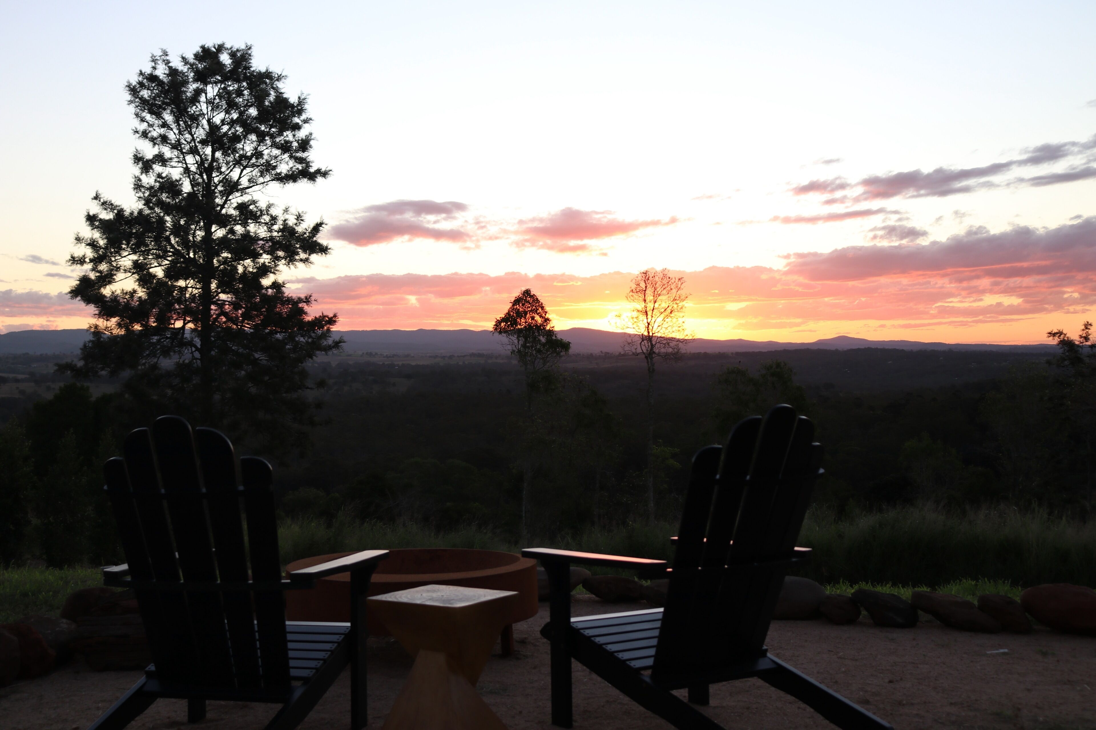 Two Adirondack chairs facing a sunset over a mountainous landscape.