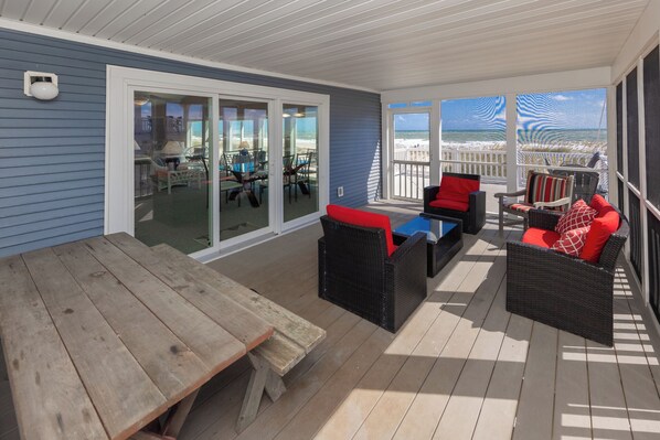 Un espacio al aire libre cubierto con una mesa de madera, muebles de mimbre con cojines rojos y vista a la playa a través de amplios ventanales.