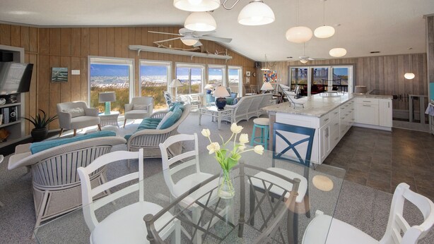 A spacious living area with a glass dining table, white wicker furniture, and a view of the ocean through large windows. - Staud's Stilts - Oceanfront home w/pool and direct beach access (Virginia Beach)