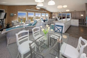Amplia sala de estar con mesa de vidrio, muebles de mimbre blanco y vista al océano a través de grandes ventanales.