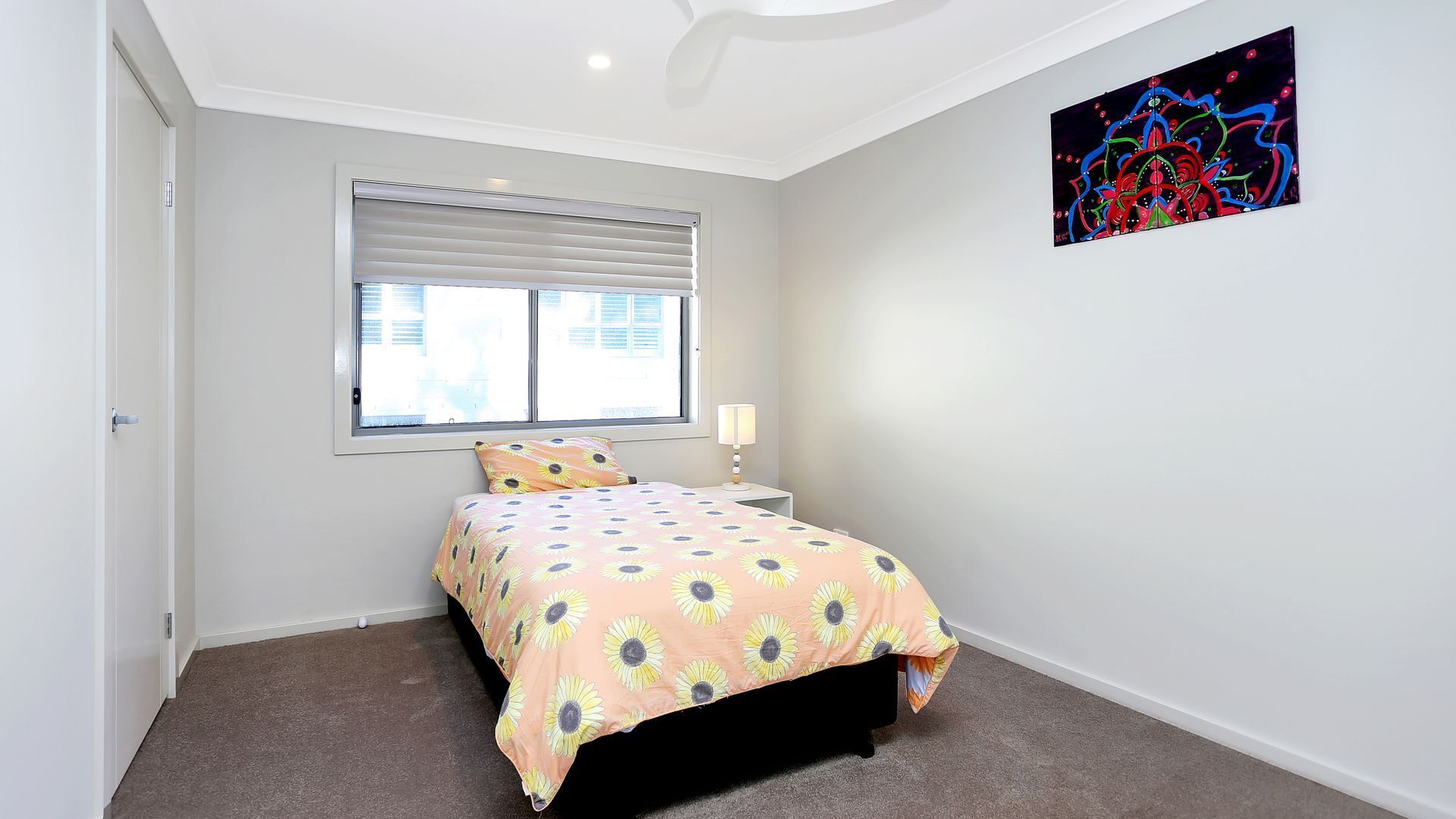 Un dormitorio con cama, ventana, mesita de noche, lámpara y una obra de arte abstracto en la pared.