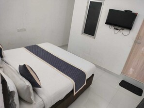 A single bed with a quilted blanket, a television mounted on the wall, and a wooden door. - Eben Service Apartment  (Chennai)