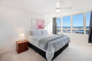 A bedroom with a bed, a nightstand, a ceiling fan, and a large window with a city view.