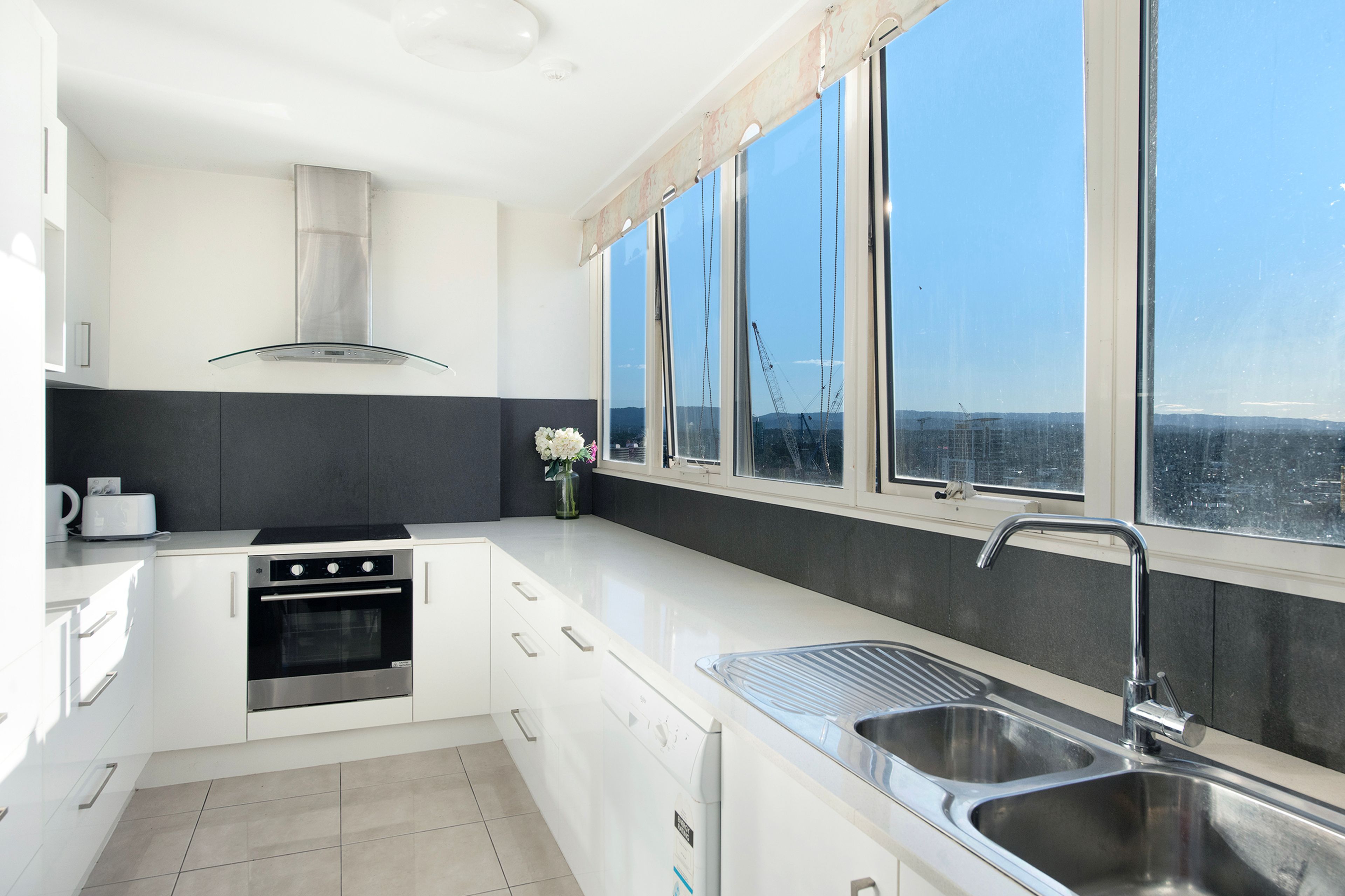 A modern kitchen with a built-in oven, a stainless steel sink, and a large window offering a view of the cityscape.
