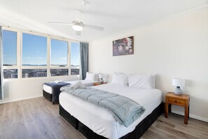 A hotel room with a large bed, a bedside table with a lamp, a window with a view of the city, and a ceiling fan.