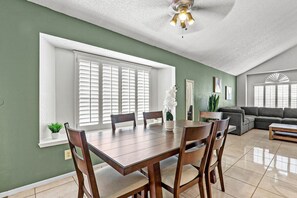 Un comedor con mesa y sillas de madera, ventilador de techo y ventanas amplias con persianas.