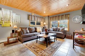 A living room with a sofa, coffee table, and two lamps. There are windows with curtains, a clock, and a bookshelf.
