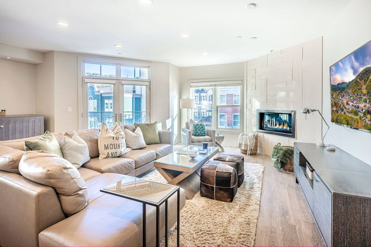 A modern living room with a sofa, coffee table, ottoman, and a flat-screen TV mounted on the wall.