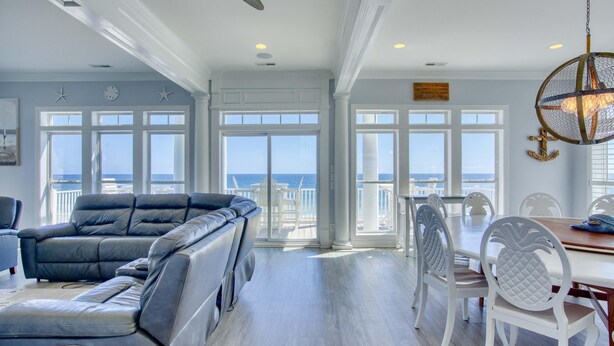 A spacious living room with a large leather sofa, white dining chairs, and a view of the ocean through large windows. - Oceanfront 7BR Sleeps 21 Hot Tub, Comm. Pool, 15 steps to the beach, 360 Views! (North Topsail Beach)