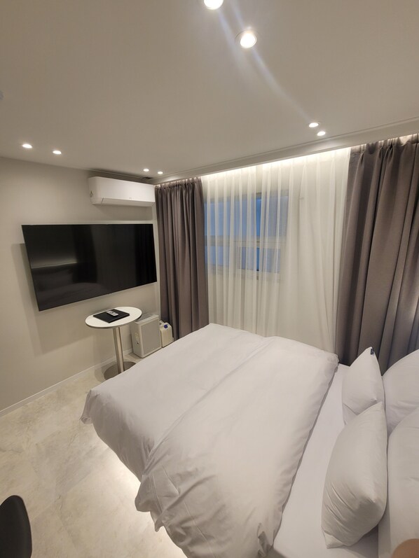 A modern hotel room with a large bed, a flat-screen TV mounted on the wall, a small round table, and a window with sheer curtains. - Hwagok August (Seoul)