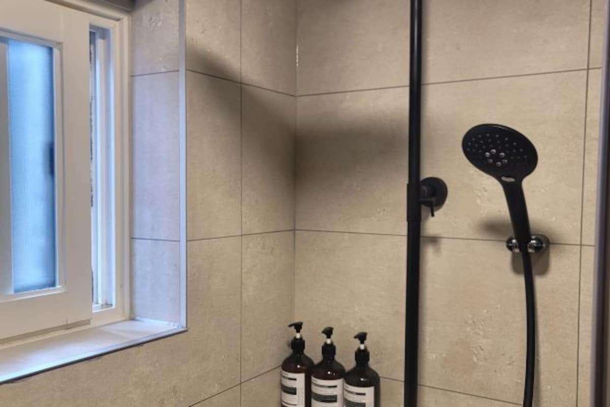 A bathroom with beige tiles, a shower with a handheld showerhead, and three bottles of liquid soap on the floor.