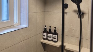 A shower with a handheld showerhead and a wall-mounted showerhead, a window, and a shelf with three bottles. - Hwagok August (Seoul)