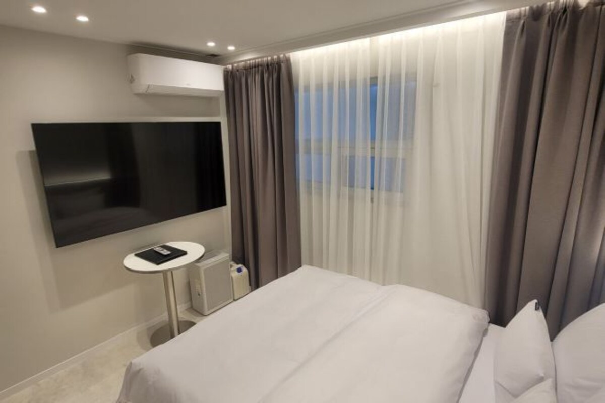 A hotel room with a bed, a TV mounted on the wall, a small table, and a window with curtains.