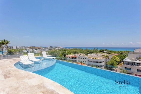 A rooftop infinity pool with white lounge chairs, surrounded by buildings and a clear view of the ocean.