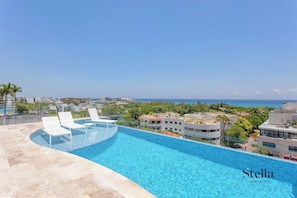 A rooftop infinity pool with white lounge chairs, surrounded by buildings and a clear view of the ocean.