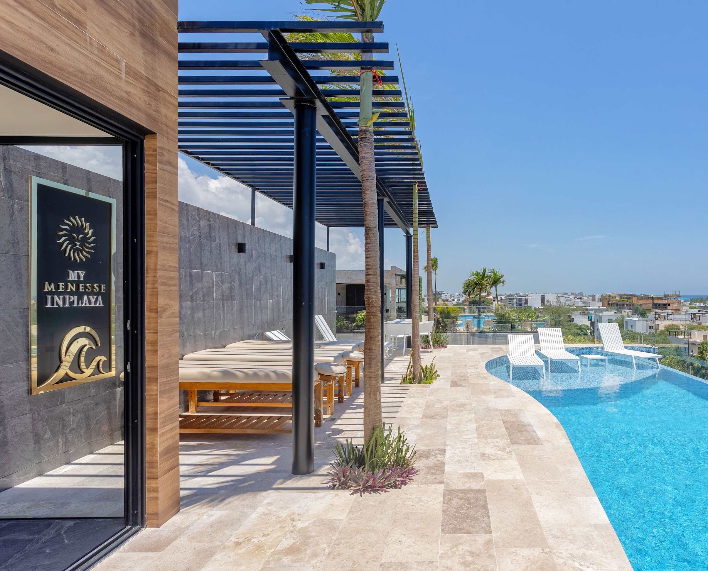 A modern poolside area with lounge chairs, a clear view of the ocean, and a sign reading 'MY MENESSE INPLAYA'.