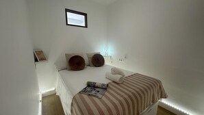 Un dormitorio minimalista con una cama, dos almohadas, una mesita de noche con una lámpara y un pequeño marco de fotos en la pared.