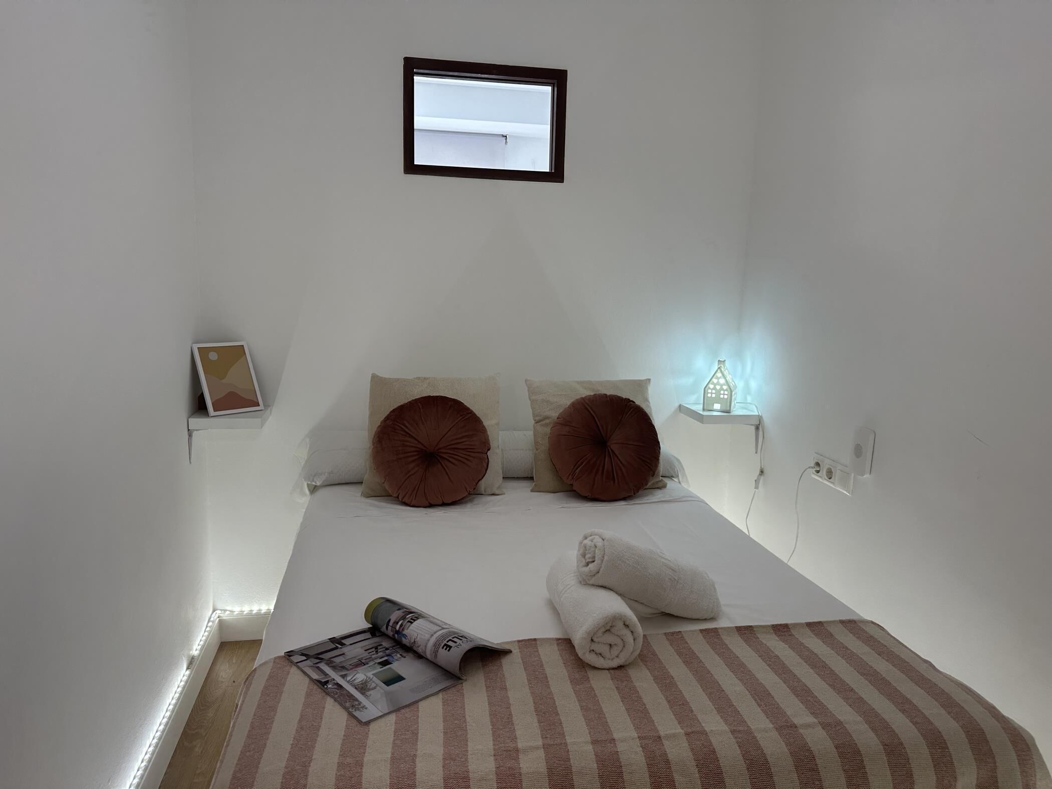 A bedroom with a bed, pillows, a bedside table with a lamp, a framed picture, and a rolled towel.