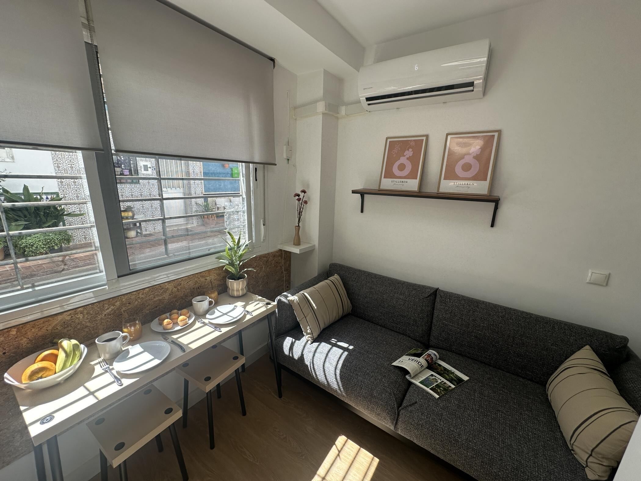 Een gezellig, compact leefruimte met een kitchenette, een eettafel met ontbijtbenodigdheden, een bank en een klein plankje met ingekaderde foto's.