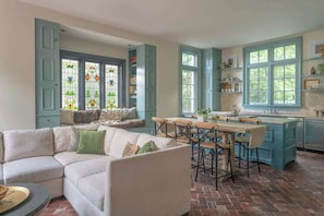 A cozy living room with a white sofa, a dining table with chairs, and a kitchen area with blue cabinets and large windows. - High Street Inn W High St Room (Charlottesville)