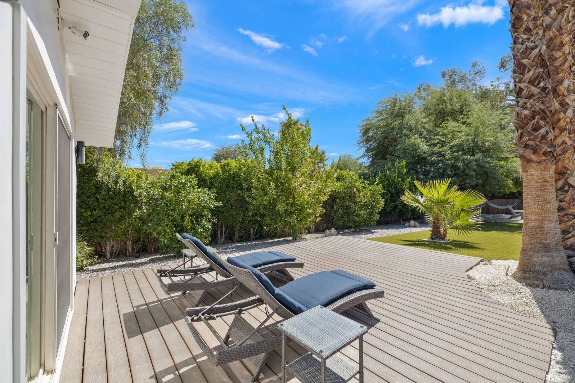 A wooden deck with lounge chairs and a small table, surrounded by greenery and a palm tree.
