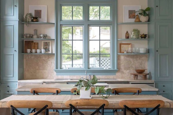 A kitchen with a large window, wooden chairs, and shelves with various items.