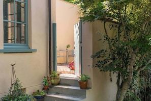 A narrow outdoor corridor with steps leading to a patio area, potted plants, and a tree.