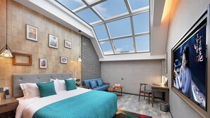A modern bedroom with a skylight, a large bed with teal bedding, a blue sofa, a small table, and a TV on the wall.